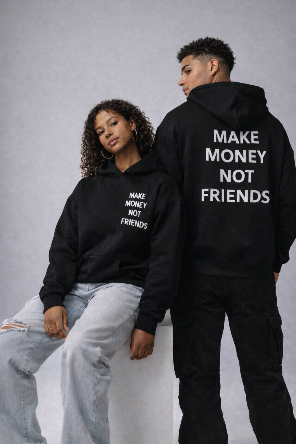 Two people wearing black hoodies with text, sitting on a gray background