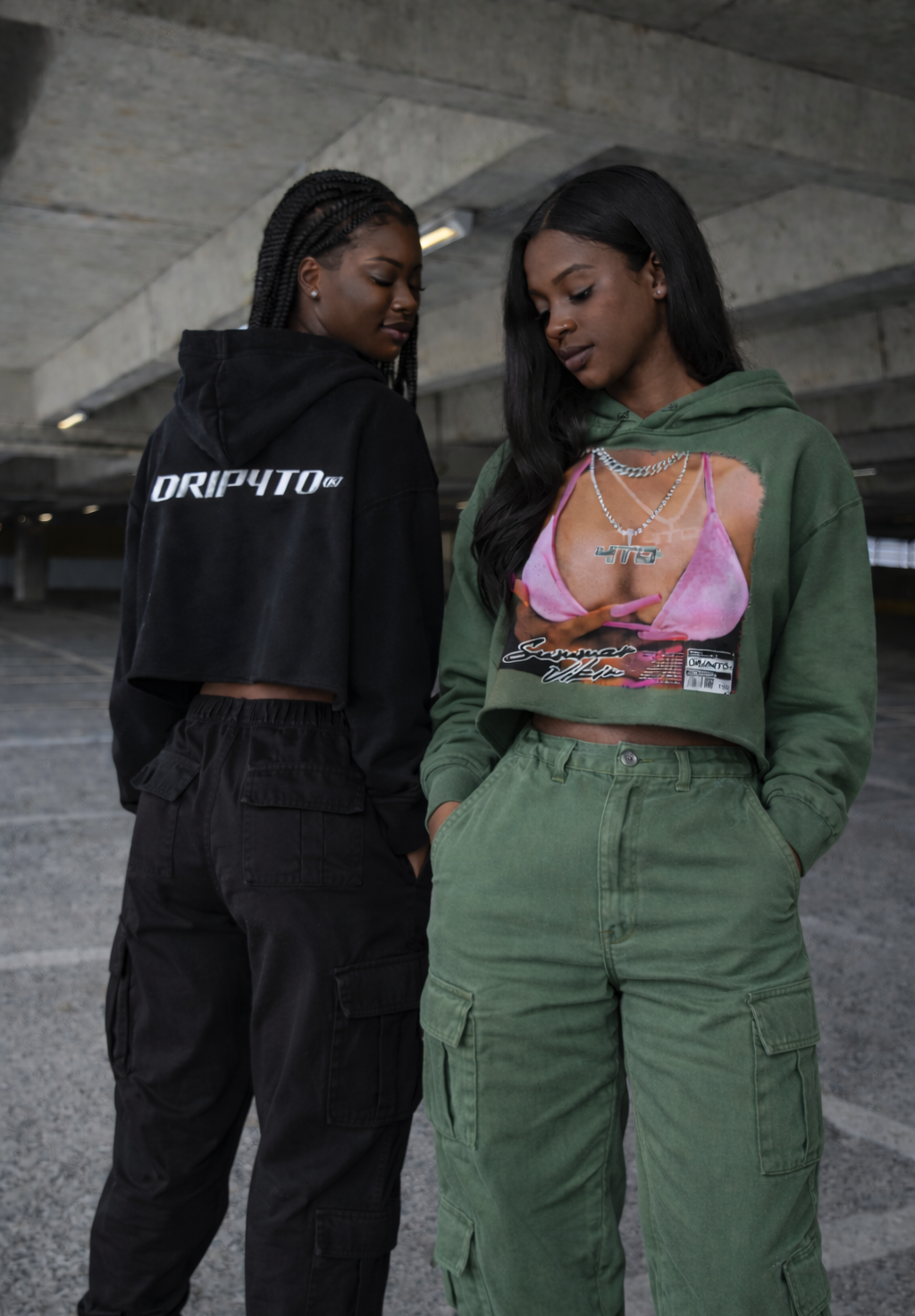Two women standing in an urban setting, one wearing a black hoodie with 'DRIP4TO' text and the other in a green hoodie with a graphic design.